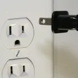 Zalane gniazdko elektryczne. Czarny wtyk zbliża się do białej obudowy, grożąc zwarciem.