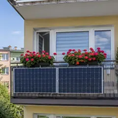 Dwa panele fotowoltaiczne na tarasie, nad nimi skrzynki z czerwonymi pelargoniami. Zielone drzewa i kolorowe budynki w tle.