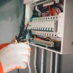 Elektryk w białych rękawicach pracuje przy skrzynce z bezpiecznikami. Widać przewody i narzędzia. Może to być związane z objawami uszkodzenia bezpiecznika automatycznego.