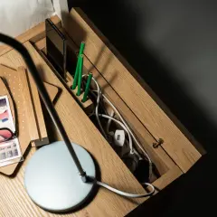 Drewniana szafka nocna z przegródkami na telefon i zielone ołówki. W schowku na kable panuje porządek dzięki praktycznym zabezpieczeniom na kable.