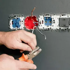 Ręce majsterkowicza przygotowują przewody do podłączenia kilku gniazdek elektrycznych. Narzędzia i instalacja na szarej ścianie.
