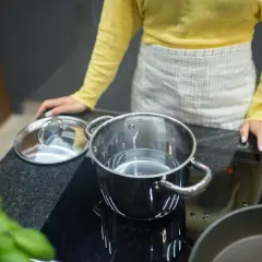 Kobieta w fartuchu przygotowuje się do gotowania na płycie indukcyjnej. W garnku jest woda, a obok leży pokrywka. Protokół podłączenia płyty indukcyjnej jest kluczowy dla bezpieczeństwa.