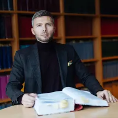 Mężczyzna w garniturze siedzi przy stole z otwartą księgą, otoczony regałami pełnymi książek. To jego zabezpieczenie STB.