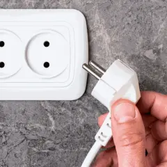 Dłoń trzyma wtyczkę do podłączenia pralki. Gniazdko elektryczne znajduje się na ścianie.