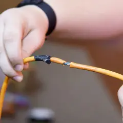 Dłoń pokazuje uszkodzony kabel pomarańczowy, gdzie widać niebieskie i białe żyłki. Pokazuje to, jak połączyć drut z linka, używając taśmy izolacyjnej.