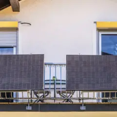 Dwa panele fotowoltaika balkonowa na balustradzie, obok okien z roletami i kwitnących donic.