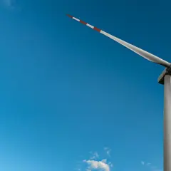 Wiatrak na tle błękitnego nieba z chmurami. Jego łopaty, ozdobione czerwono-białymi pasami, obracają się, czerpiąc czystą energię z wiatru.