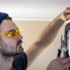 Elektryk w okularach ochronnych i czapce podłącza przewody w puszce instalacyjnej, trzymając kombinerki.
