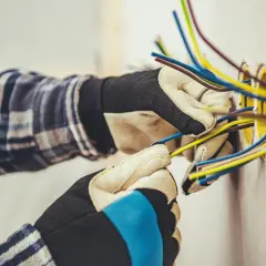 Elektryk w rękawicach przygotowuje przewody do podłączenia gniazdek. Widać kilka kolorowych kabli wychodzących ze ściany, gotowych do połączenia. To kluczowy etap, jak zaznaczyć gniazdka na projekcie.