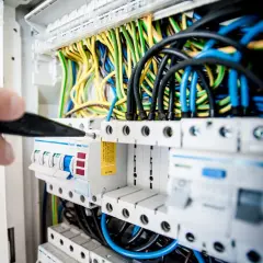Elektryk naprawia awarię instalacji elektrycznej, manipulując śrubokrętem przy bezpiecznikach i plątaninie kolorowych przewodów.