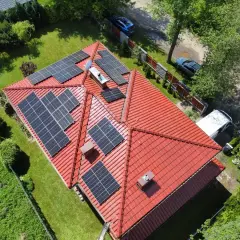 Panele fotowoltaiczne na czerwonym dachu, instalacja wykonana na starych zasadach. Wokół zieleń i samochody.