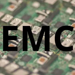 Na zielonej płytce drukowanej z widocznymi elementami elektronicznymi, wielkimi czarnymi literami widnieje napis "EMC". To skrót od Electromagnetic Compatibility, czyli kompatybilności elektromagnetycznej.