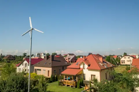 Biała turbina wiatrowa na tle błękitnego nieba i domów z czerwonymi dachami.