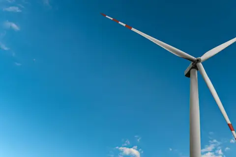 Wiatrak na tle błękitnego nieba z chmurami. Jego łopaty, ozdobione czerwono-białymi pasami, obracają się, czerpiąc czystą energię z wiatru.