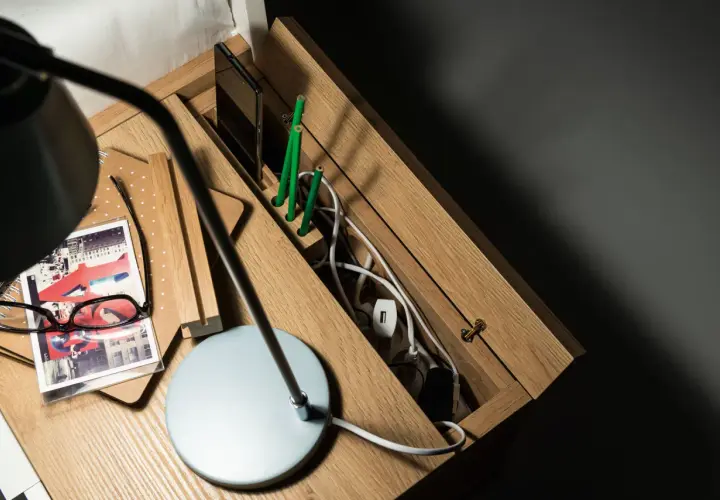 Drewniana szafka nocna z przegródkami na telefon i zielone ołówki. W schowku na kable panuje porządek dzięki praktycznym zabezpieczeniom na kable.