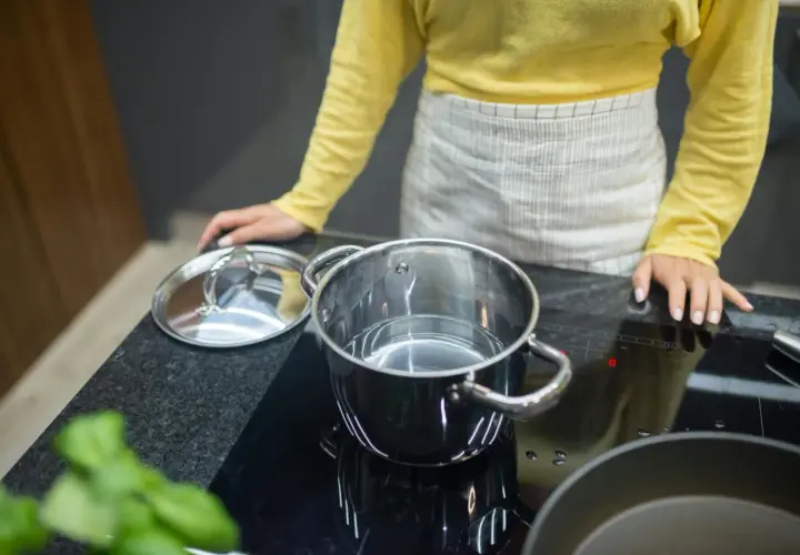 Kobieta w fartuchu przygotowuje się do gotowania na płycie indukcyjnej. W garnku jest woda, a obok leży pokrywka. Protokół podłączenia płyty indukcyjnej jest kluczowy dla bezpieczeństwa.