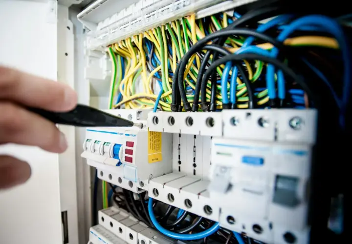 Ręka z narzędziem przyłącze trójfazowe. Widać bezpieczniki i plątaninę kolorowych przewodów w rozdzielnicy elektrycznej.