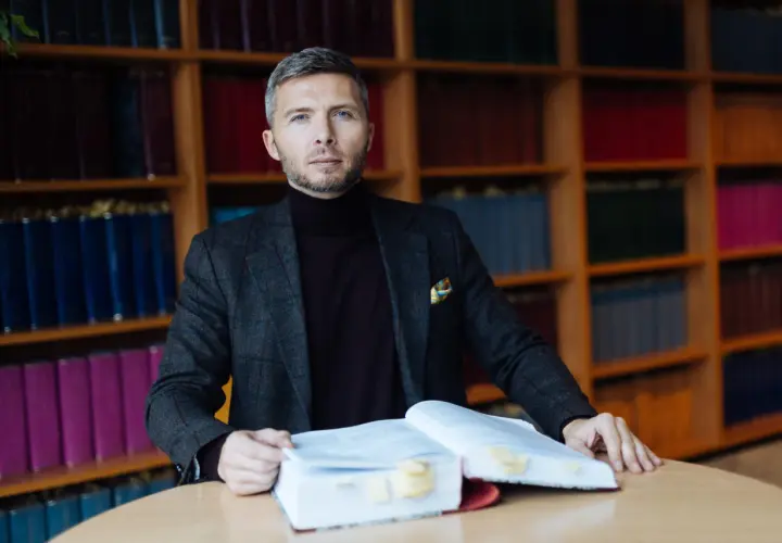 Mężczyzna w garniturze siedzi przy stole z otwartą księgą, otoczony regałami pełnymi książek. To jego zabezpieczenie STB.