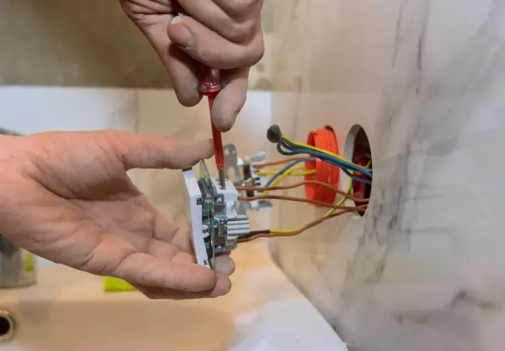 Ręce majsterkowicza montują gniazdko, przykręcając śrubki. W ścianie widać kolorowe przewody elektryczne, gotowe do podłączenia.
