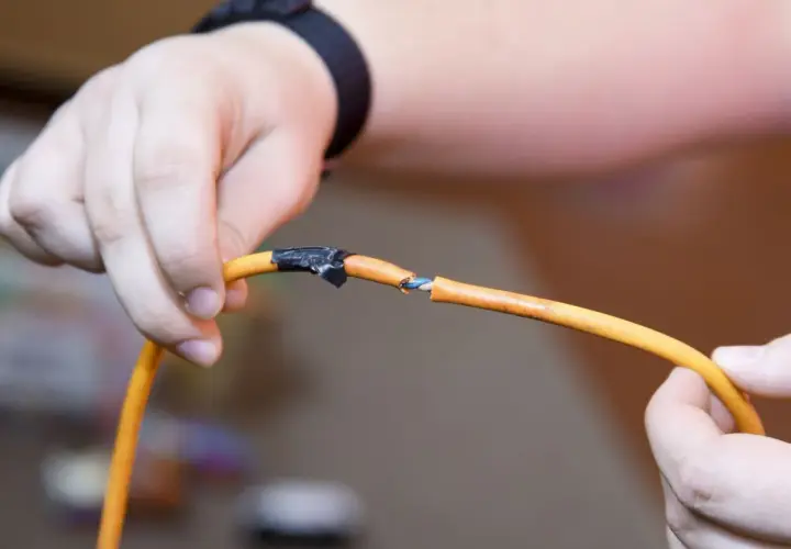 Dłoń pokazuje uszkodzony kabel pomarańczowy, gdzie widać niebieskie i białe żyłki. Pokazuje to, jak połączyć drut z linka, używając taśmy izolacyjnej.