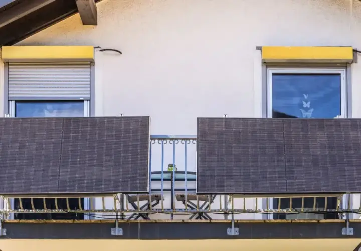 Dwa panele fotowoltaika balkonowa na balustradzie, obok okien z roletami i kwitnących donic.