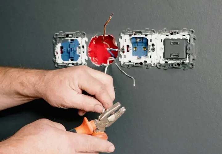 Elektryk łączy przewody, przygotowując instalację, jak podłączyć 3 gniazda obok siebie. Widać pęsetę i odsłonięte kable.