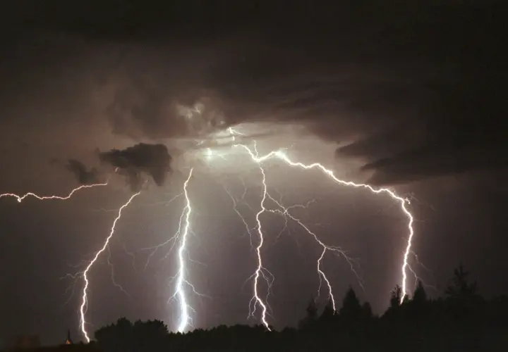 Potężne wyładowania elektryczne rozświetlają nocne niebo, ukazując, ile volt ma piorun. Burza nad horyzontem z drzewami w ciemnej sylwetce.