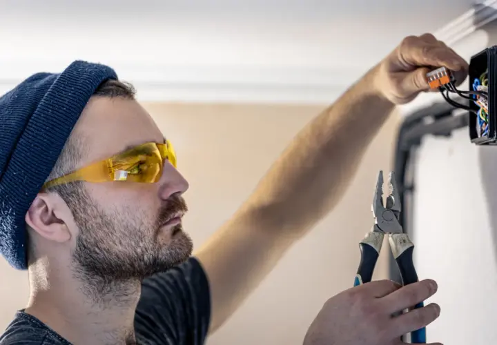 Elektryk w okularach ochronnych i czapce podłącza przewody w puszce instalacyjnej, trzymając kombinerki.