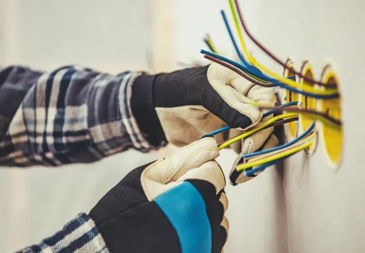 Elektryk w rękawicach przygotowuje przewody do podłączenia gniazdek. Widać kilka kolorowych kabli wychodzących ze ściany, gotowych do połączenia. To kluczowy etap, jak zaznaczyć gniazdka na projekcie.