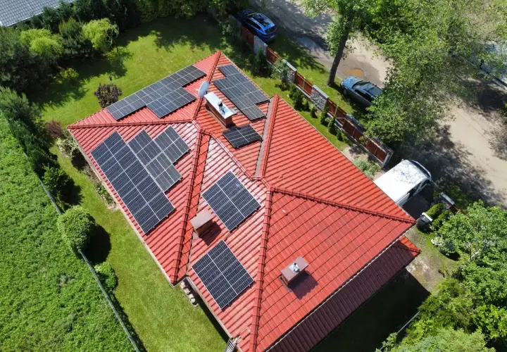Panele fotowoltaiczne na czerwonym dachu, instalacja wykonana na starych zasadach. Wokół zieleń i samochody.