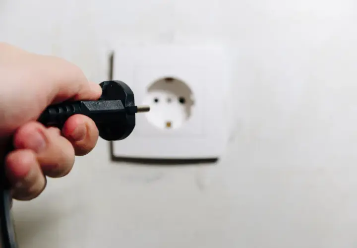 Dłoń trzyma wtyczkę, zbliżając ją do gniazdka. Uważaj, by nie doświadczyć porażenie prądem z gniazdka!