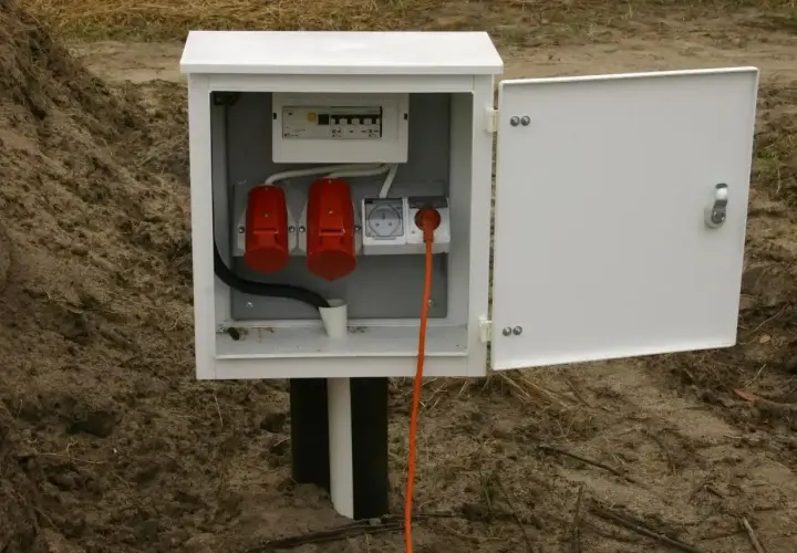 Biała skrzynka z bezpiecznikami i gniazdami, stanowiąca przyłącze elektryczne do domu. Pomarańczowy kabel wychodzi z gniazdka.