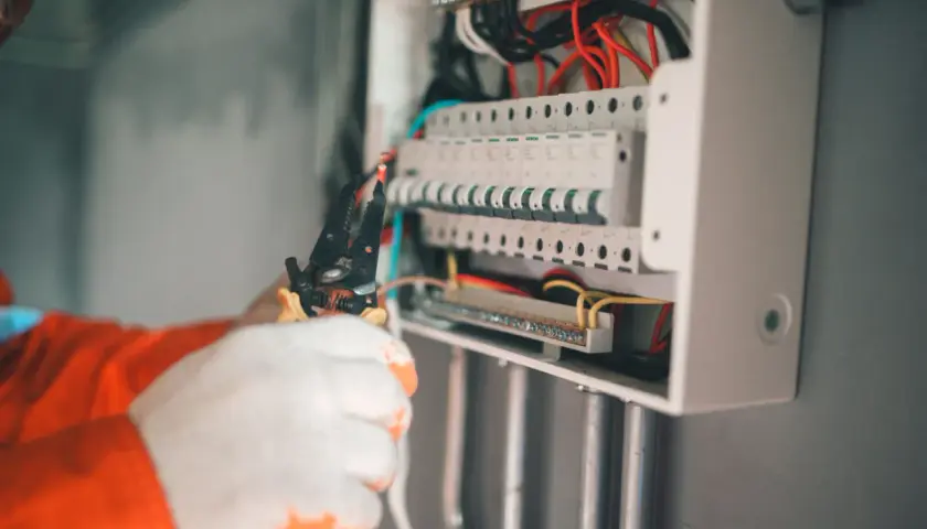 Elektryk w białych rękawicach pracuje przy skrzynce z bezpiecznikami. Widać przewody i narzędzia. Może to być związane z objawami uszkodzenia bezpiecznika automatycznego.