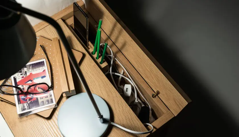 Drewniana szafka nocna z przegródkami na telefon i zielone ołówki. W schowku na kable panuje porządek dzięki praktycznym zabezpieczeniom na kable.