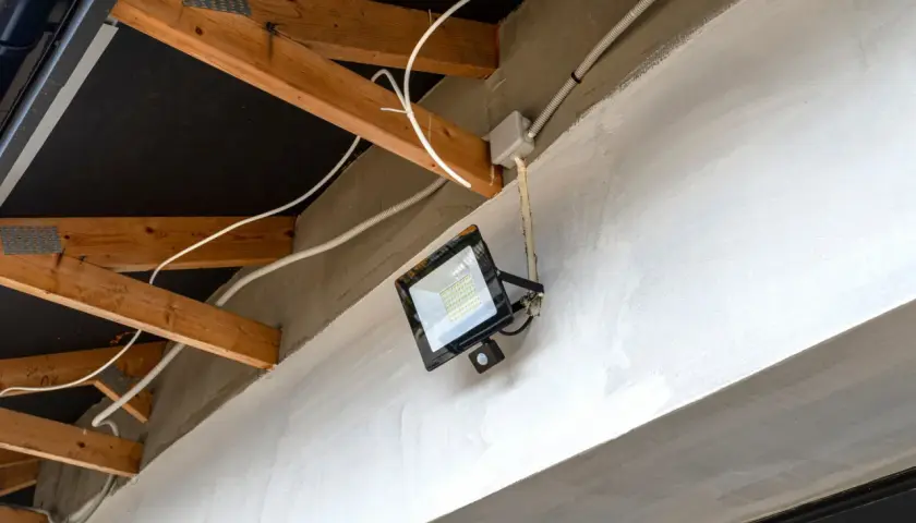 Nowoczesna elektryka w garażu: jasny reflektor LED z czujnikiem ruchu zamontowany na betonowej belce, otoczony drewnianą konstrukcją dachu i białymi przewodami.