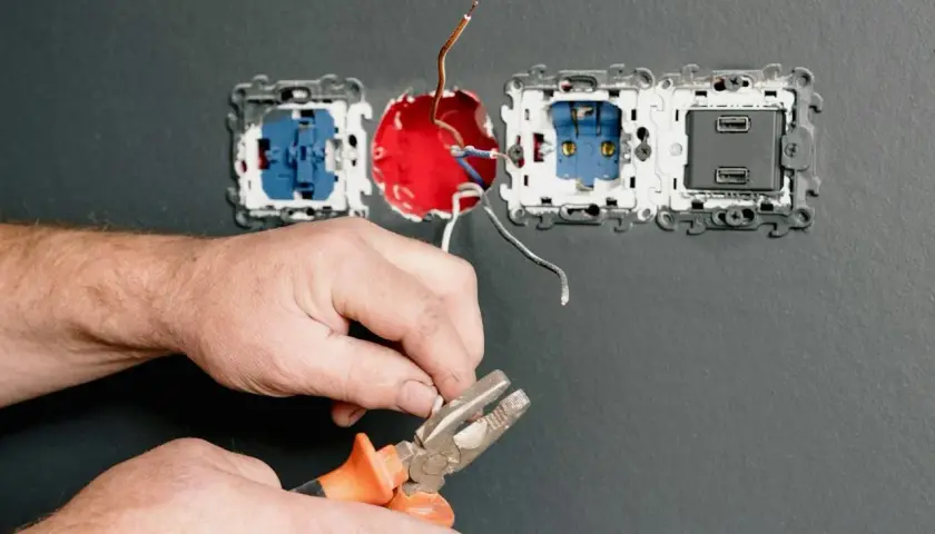 Ręce majsterkowicza przygotowują przewody do podłączenia kilku gniazdek elektrycznych. Narzędzia i instalacja na szarej ścianie.