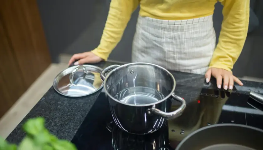 Kobieta w fartuchu przygotowuje się do gotowania na płycie indukcyjnej. W garnku jest woda, a obok leży pokrywka. Protokół podłączenia płyty indukcyjnej jest kluczowy dla bezpieczeństwa.