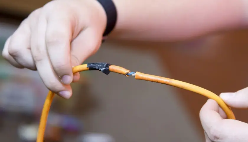 Dłoń pokazuje uszkodzony kabel pomarańczowy, gdzie widać niebieskie i białe żyłki. Pokazuje to, jak połączyć drut z linka, używając taśmy izolacyjnej.