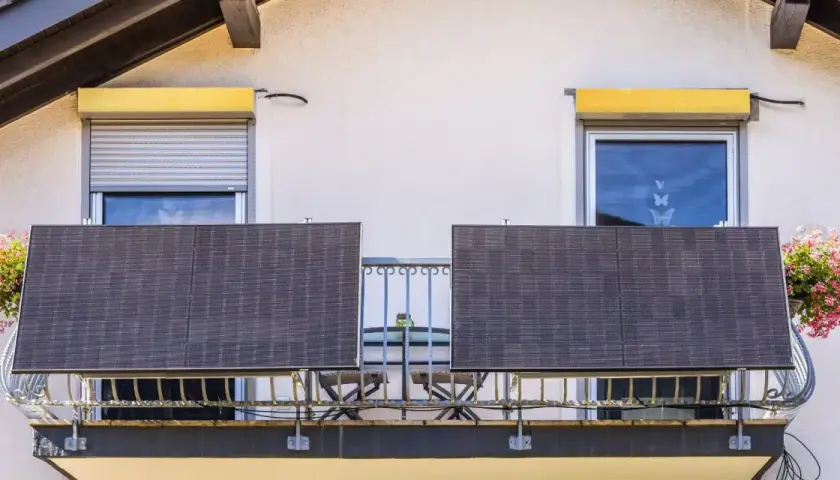 Dwa panele fotowoltaika balkonowa na balustradzie, obok okien z roletami i kwitnących donic.