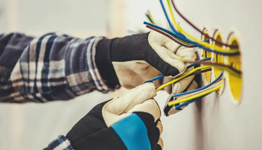 Elektryk w rękawicach przygotowuje przewody do podłączenia gniazdek. Widać kilka kolorowych kabli wychodzących ze ściany, gotowych do połączenia. To kluczowy etap, jak zaznaczyć gniazdka na projekcie.