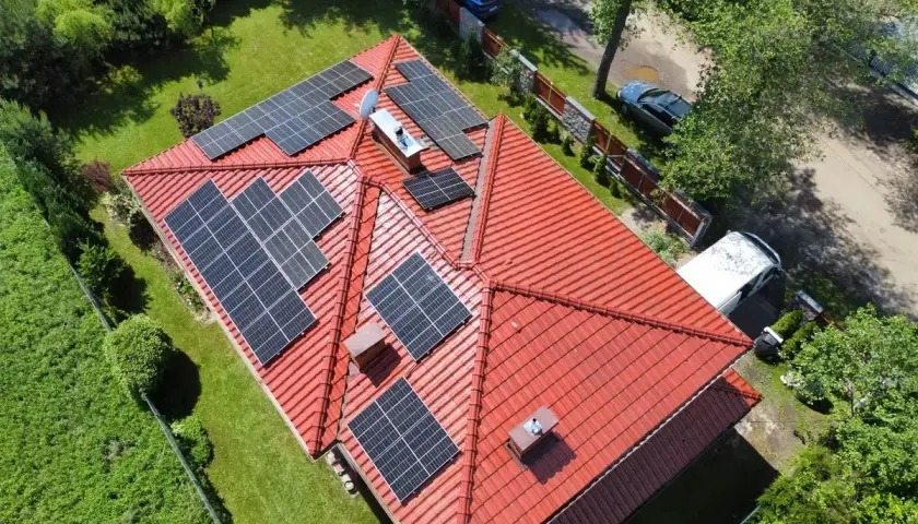 Panele fotowoltaiczne na czerwonym dachu, instalacja wykonana na starych zasadach. Wokół zieleń i samochody.