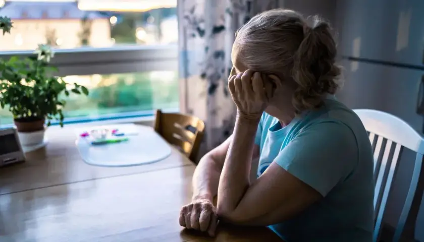Starsza kobieta siedzi przy stole, zmartwiona. Zima i odcięcie prądu sprawiają, że czuje się samotna i bezradna.
