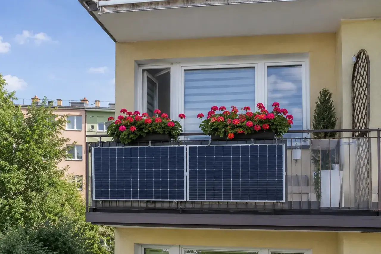 Dwa panele fotowoltaiczne na tarasie, nad nimi skrzynki z czerwonymi pelargoniami. Zielone drzewa i kolorowe budynki w tle.
