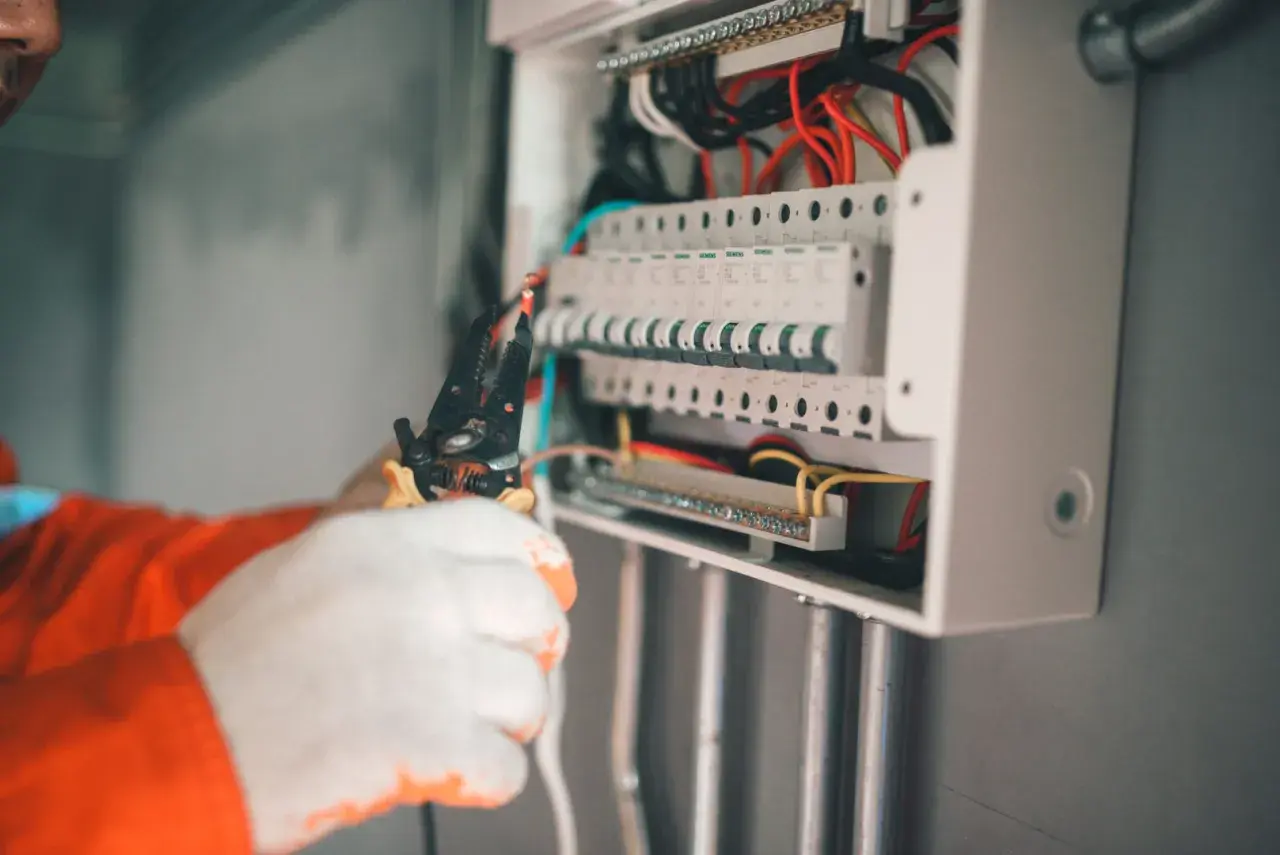 Elektryk w białych rękawicach pracuje przy skrzynce z bezpiecznikami. Widać przewody i narzędzia. Może to być związane z objawami uszkodzenia bezpiecznika automatycznego.