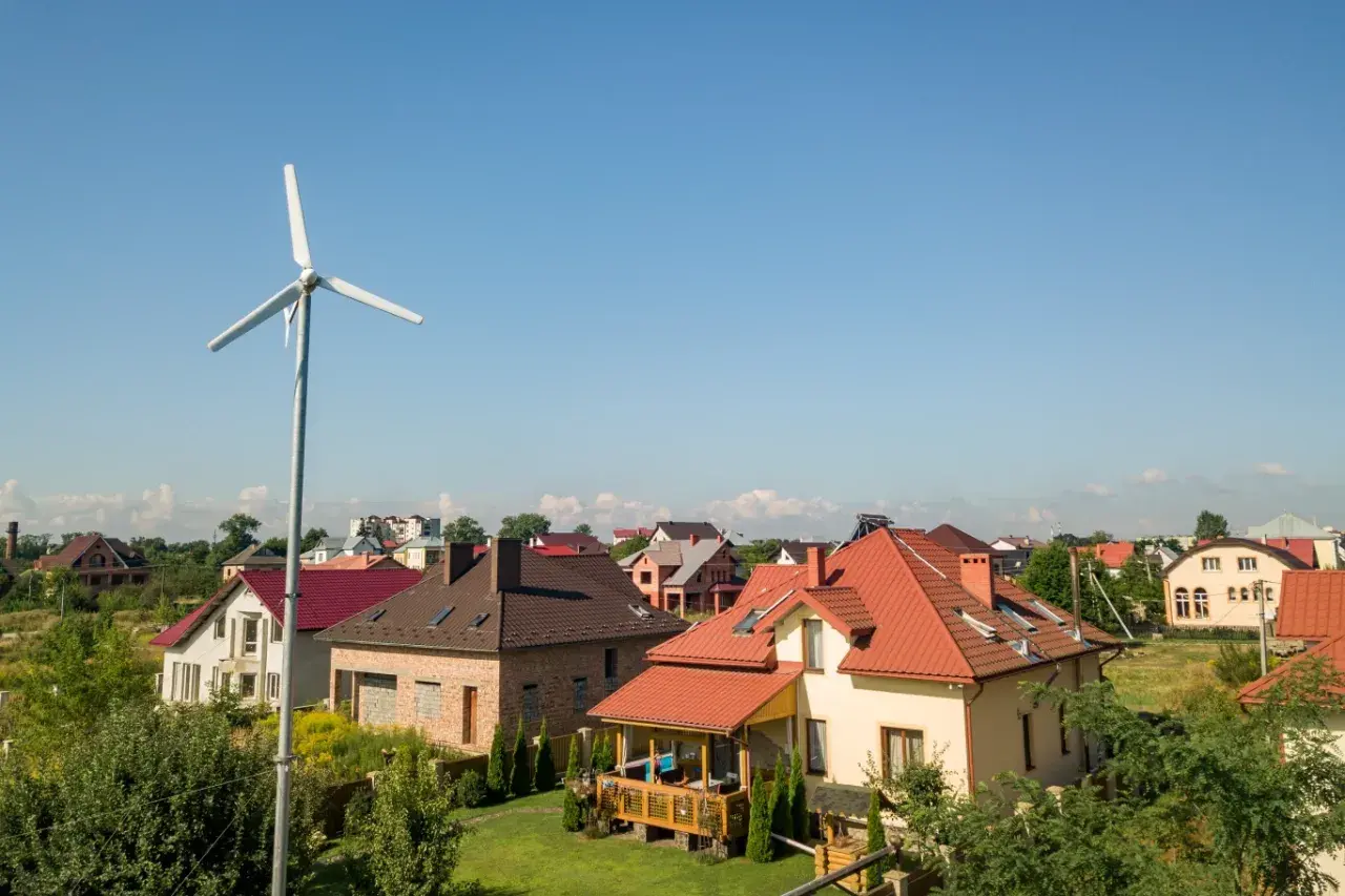 Biała turbina wiatrowa na tle błękitnego nieba i domów z czerwonymi dachami.