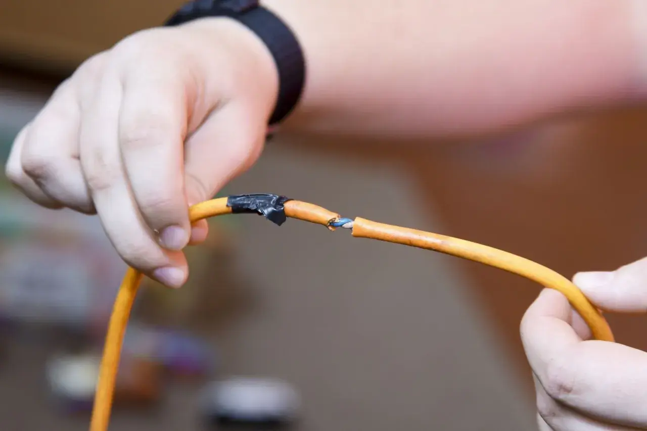 Dłoń pokazuje uszkodzony kabel pomarańczowy, gdzie widać niebieskie i białe żyłki. Pokazuje to, jak połączyć drut z linka, używając taśmy izolacyjnej.