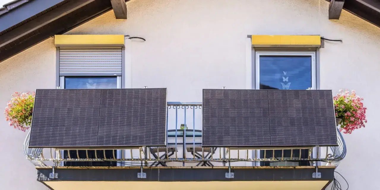 Dwa panele fotowoltaika balkonowa na balustradzie, obok okien z roletami i kwitnących donic.