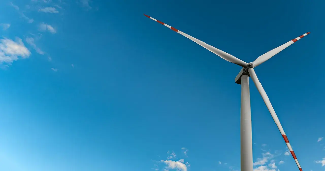 Wiatrak na tle błękitnego nieba z chmurami. Jego łopaty, ozdobione czerwono-białymi pasami, obracają się, czerpiąc czystą energię z wiatru.