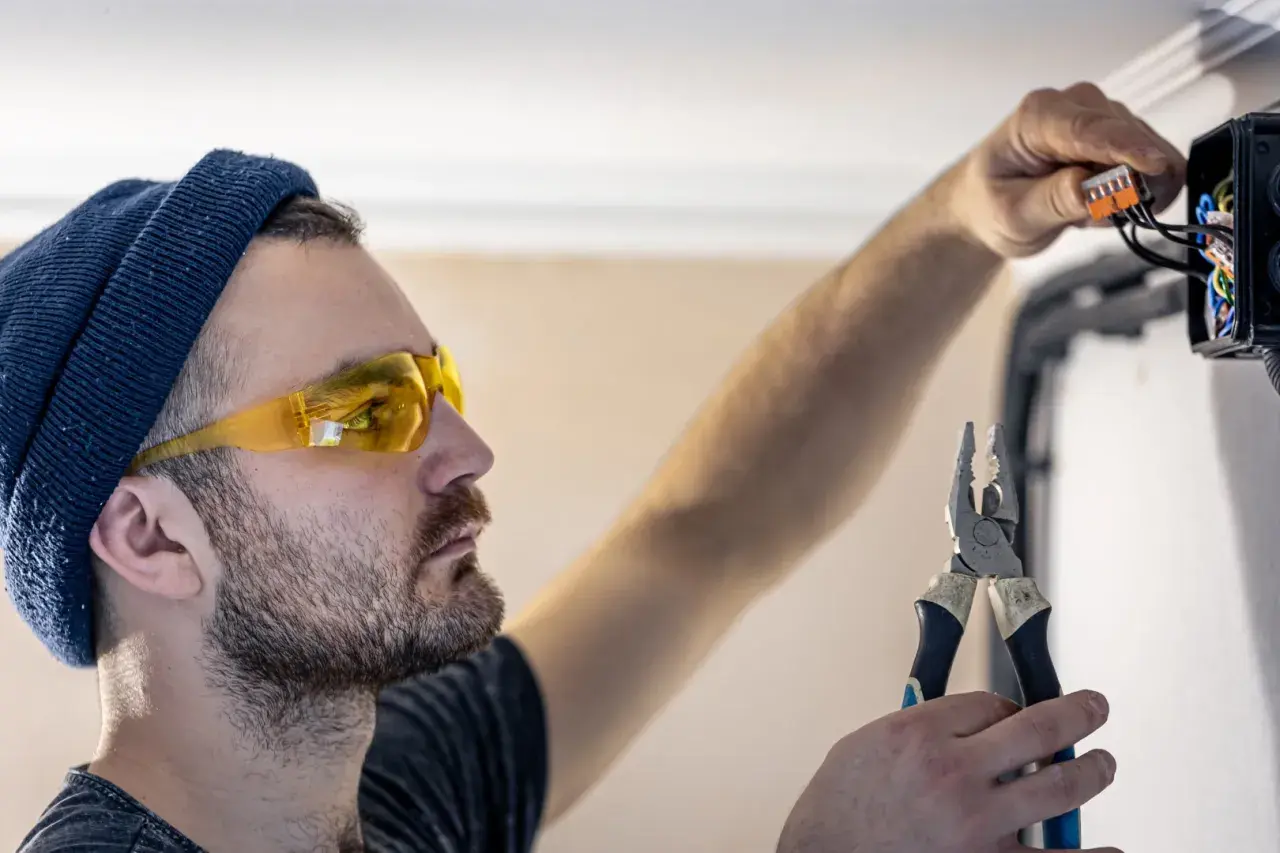 Elektryk w okularach ochronnych i czapce podłącza przewody w puszce instalacyjnej, trzymając kombinerki.