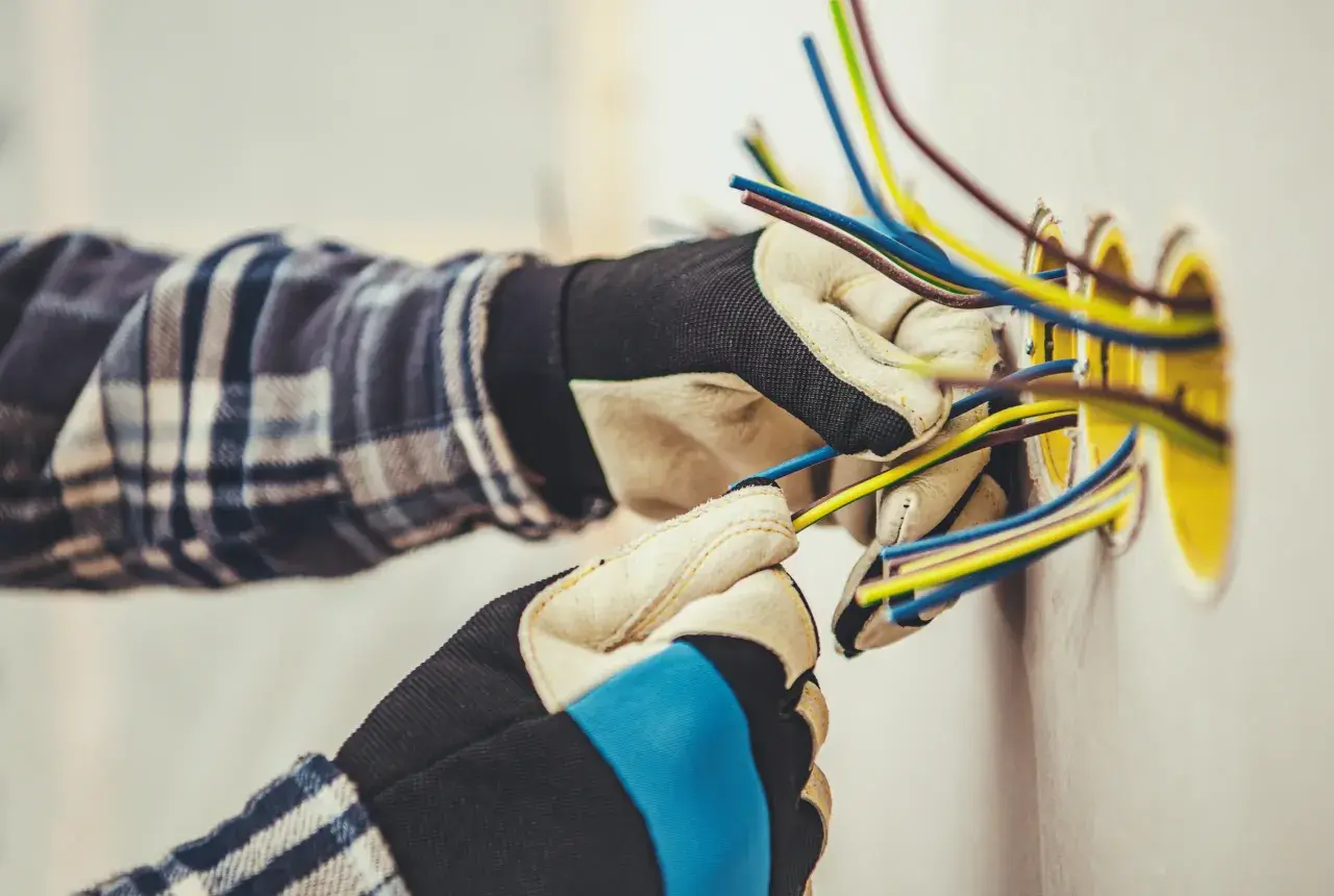Elektryk w rękawicach przygotowuje przewody do podłączenia gniazdek. Widać kilka kolorowych kabli wychodzących ze ściany, gotowych do połączenia. To kluczowy etap, jak zaznaczyć gniazdka na projekcie.